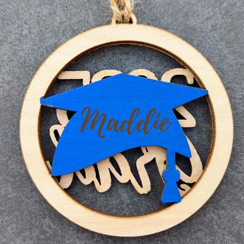 May include: A wooden round ornament with a blue graduation cap cutout. The cap has the name "Maddie" written on it. The ornament has the words "Class of 2023" cut out around the cap.