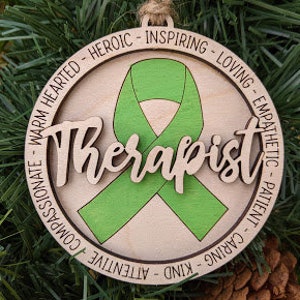 May include: Wooden ornament with a green ribbon and the word "Therapist" in a decorative font. The ornament is surrounded by a circle of words describing positive qualities, such as "warm hearted", "heroic", "inspiring", "loving", "empathetic", "patient", "caring", "kind", "attentive", and "compassionate".