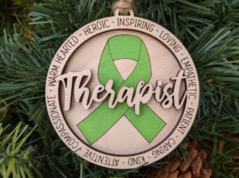 May include: Wooden ornament with a green ribbon and the word "Therapist" in a decorative font. The ornament is surrounded by a circle of words describing positive qualities, such as "warm hearted", "heroic", "inspiring", "loving", "empathetic", "patient", "caring", "kind", "attentive", and "compassionate".
