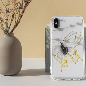 以下が含まれることがあります： 水彩の蝶のデザインが施された透明な携帯電話ケース。蝶は黄色いアクセントのある白で、右に飛んでいます。ケースは白い表面にあります。