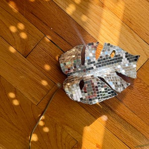 May include: A decorative monstera leaf ornament covered in small, square, reflective silver mirror tiles. The leaf shape features characteristic cutouts and is attached to a thin, flexible wire stem, creating a mosaic effect that catches light.
