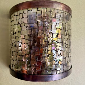 May include: A semi-cylindrical wall sconce featuring a mosaic of iridescent, reflective glass tiles arranged in a grid pattern. The fixture is framed by a smooth, dark copper-toned metal band at both the top and bottom edges, creating a contrast.