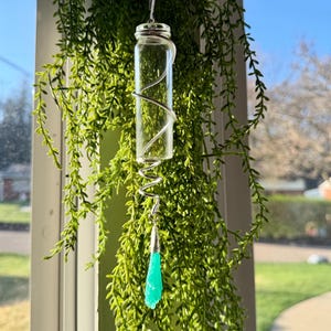 Puede incluir: Un vial de vidrio transparente envuelto con alambre plateado, suspendido por una cadena plateada. Un cristal verde claro en forma de lágrima cuelga debajo. El vial está rodeado de plantas verdes colgantes, con una ventana brillante en el fondo.