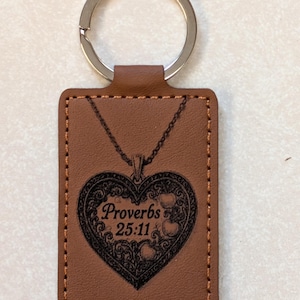 May include: A brown leather keychain with a silver ring. The rectangular tag features a heart-shaped pendant design with the text "Proverbs 25:11" in black. The edges are stitched with a lighter brown thread.