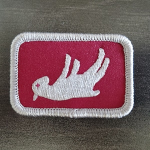 Puede incluir: Un parche bordado blanco con una imagen estilizada de un gato sobre un fondo rojo. El parche tiene un borde blanco.