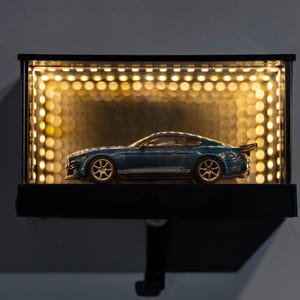 Pode incluir: Um carro de brinquedo azul com aros dourados é exibido dentro de uma vitrine preta iluminada. A vitrine é iluminada por pequenas luzes de tons quentes, criando um efeito de holofote. O carro tem um spoiler.