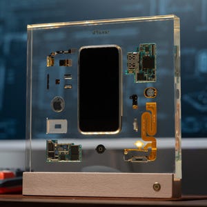 以下が含まれることがあります： 透明なアクリルブロックに収められたiPhone。内部コンポーネントが展示されています。黒い画面が中央にあり、基板、ケーブル、Appleのロゴが周囲に配置されています。ベースは明るい色の木材です。