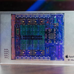 以下が含まれることがあります： Apple M1 Maxチップの詳細な画像。複雑な内部コンポーネントを示しています。チップは透明な長方形のブロックに収められています。デザインは、Appleのロゴとチップの仕様とともに、回路とプロセッサの複雑な配置を示しています。
