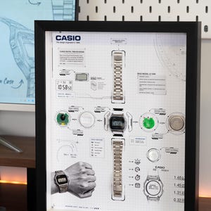 May include: A black framed wall art display featuring an exploded view of a Casio A158W digital watch. The print includes technical diagrams, component labels, a wrist-worn watch illustration, and the Casio logo on a white grid background.