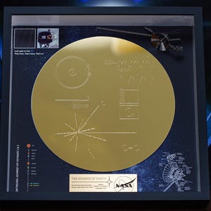 May include: Framed gold-coloured Voyager Golden Record replica. The record features etched diagrams and symbols, including a star map and scientific data. The frame includes text and a NASA logo, with a dark blue background.