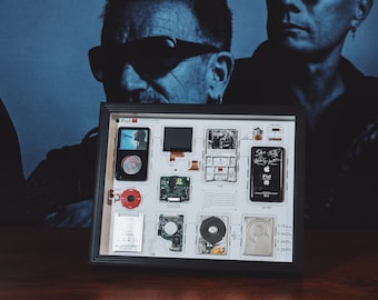 May include: A framed display of the components of an Apple iPod, including the original iPod, circuit boards, hard drives, and a signed note. The display is in a black frame and is on a wooden surface.