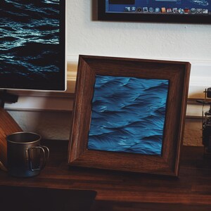 K&ouml;nnte beinhalten: Ein dunkler Holzrahmen mit einem blauen und schwarzen abstrakten Wellenmusterdruck im Inneren. Der Rahmen steht auf einem Holztisch mit einer schwarzen Tasse und einer Kamera.