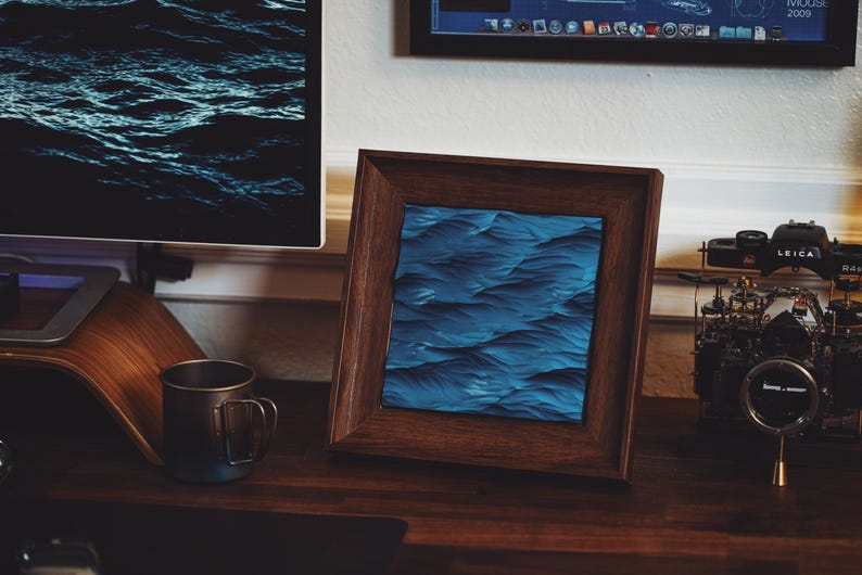 K&ouml;nnte beinhalten: Ein dunkler Holzrahmen mit einem blauen und schwarzen abstrakten Wellenmusterdruck im Inneren. Der Rahmen steht auf einem Holztisch mit einer schwarzen Tasse und einer Kamera.