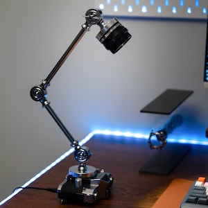 Könnte beinhalten: Eine silberne und schwarze Gelenk-Schreibtischlampe mit zylindrischem Kopf. Die Lampe hat mehrere Gelenke, die eine flexible Positionierung ermöglichen. Sie steht auf einem schwarzen Sockel und ist an eine Stromquelle angeschlossen. Der Hintergrund umfasst einen Schreibtisch mit Computer und Tastatur.