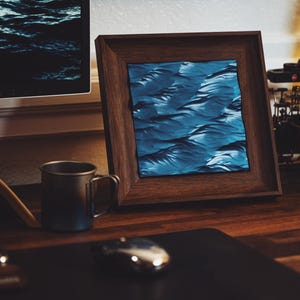 May include: A framed abstract art print with a blue and white wave pattern. The frame is made of dark wood and is sitting on a wooden desk.