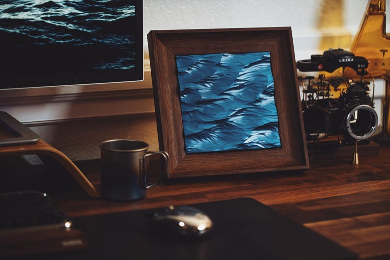 K&ouml;nnte beinhalten: Ein gerahmter abstrakter Kunstdruck mit einem blauen und wei&szlig;en Wellenmuster. Der Rahmen ist aus dunklem Holz und steht auf einem Holztisch.