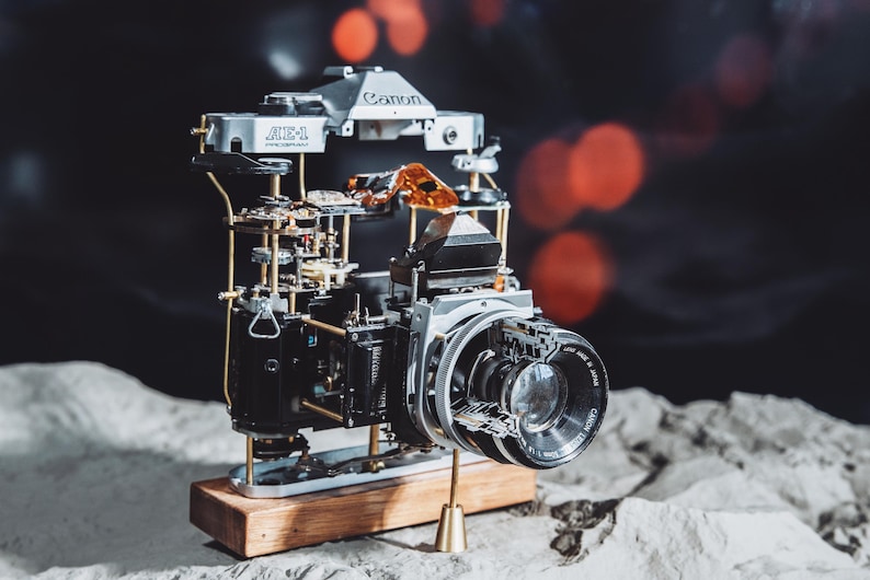 May include: A disassembled Canon AE-1 camera with a silver and black finish, mounted on a wooden base. The camera is partially exposed, revealing its internal mechanisms.