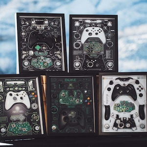 Könnte beinhalten: Gerahmte Kunstwerke mit zerlegten Xbox-Controllern. Jedes Stück zeigt die inneren Komponenten der Controller, einschließlich Leiterplatten und Tasten, in einem detaillierten, technischen Stil. Die Farbpalette umfasst Schwarz, Weiß und Grün.