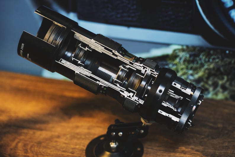 May include: A black camera lens with the internal mechanisms exposed, showing the intricate workings of the lens. The lens is mounted on a black tripod.