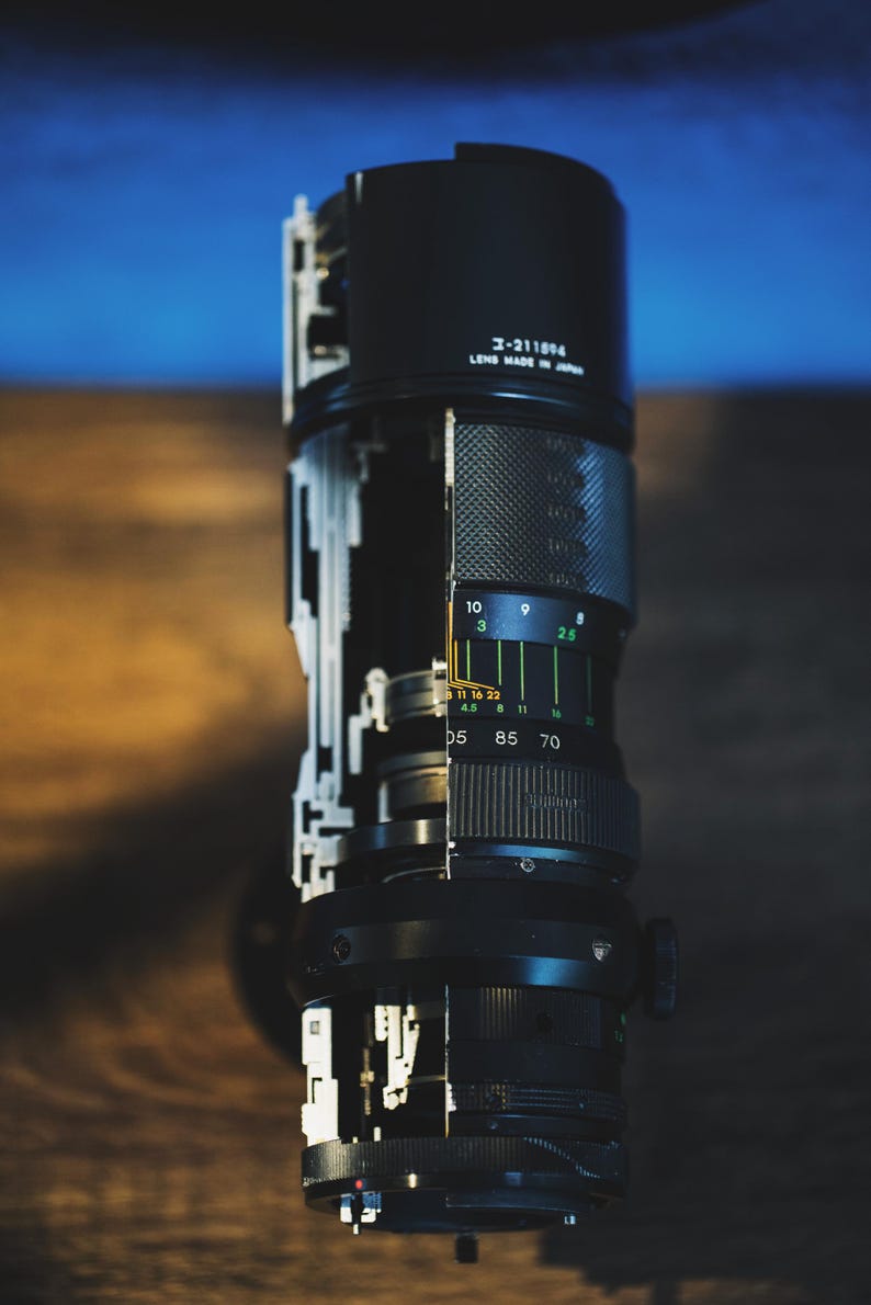 May include: A disassembled camera lens with a black exterior and silver accents. The lens has a series of rings with numbers and markings, including "コー211594" and "LENS MADE IN JAPAN".