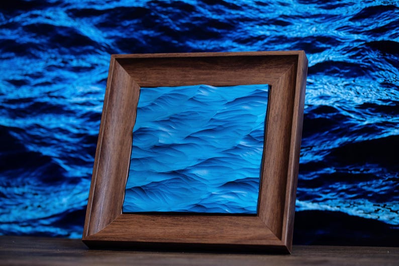 K&ouml;nnte beinhalten: Ein gerahmtes Kunstwerk, das ein blaues Meereswellenmuster darstellt. Der Rahmen ist aus dunklem Holz und hat ein schlichtes Design.