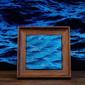 K&ouml;nnte beinhalten: Ein gerahmtes Gem&auml;lde von blauen Ozeanwellen. Das Gem&auml;lde ist in einem dunklen Holzrahmen und vor einem blauen Hintergrund platziert.