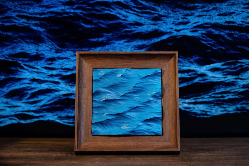 K&ouml;nnte beinhalten: Ein gerahmtes Gem&auml;lde von blauen Ozeanwellen. Das Gem&auml;lde ist in einem dunklen Holzrahmen und vor einem blauen Hintergrund platziert.