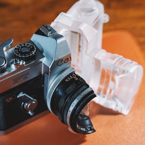 May include: A vintage Canon FT QL camera with a black lens and silver accents. The camera is positioned next to a clear plastic accessory. The camera is on a brown surface.