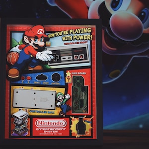 May include: A framed print of a retro video game console with a red and white color scheme. The print features a cartoon character in a red suit and hat, and the text "NOW YOU'RE PLAYING WITH POWER!" The print also includes a Nintendo Entertainment System logo.