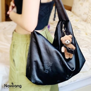 May include: Black satin tote bag with a floral pattern and a small brown teddy bear keychain. The bag is held by a person wearing a black top and green pants. The bag has a unique shape and design.