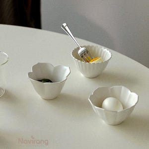 May include: Three white, petal-shaped bowls on a white surface. One bowl holds a hard-boiled egg, another has a green item, and the third contains a piece of egg with a fork. The bowls have a fluted design.