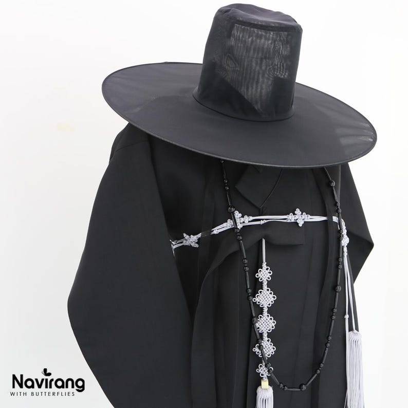May include: A black traditional Korean garment with long sleeves and a high collar, paired with a wide-brimmed black hat made of sheer material. A decorative white knotted cord and a long black beaded necklace with white tassels hang over the front.