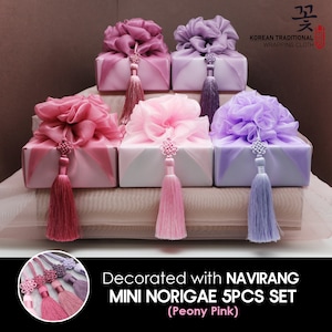May include: Five small gift boxes wrapped in traditional Korean wrapping cloth, called "Norigae", with pink, purple, and white fabric. The boxes are decorated with a flower-shaped bow and a tassel. The text "Decorated with NAVIRANG MINI NORIGAE 5PCS SET (Peony Pink)" is at the bottom of the image.