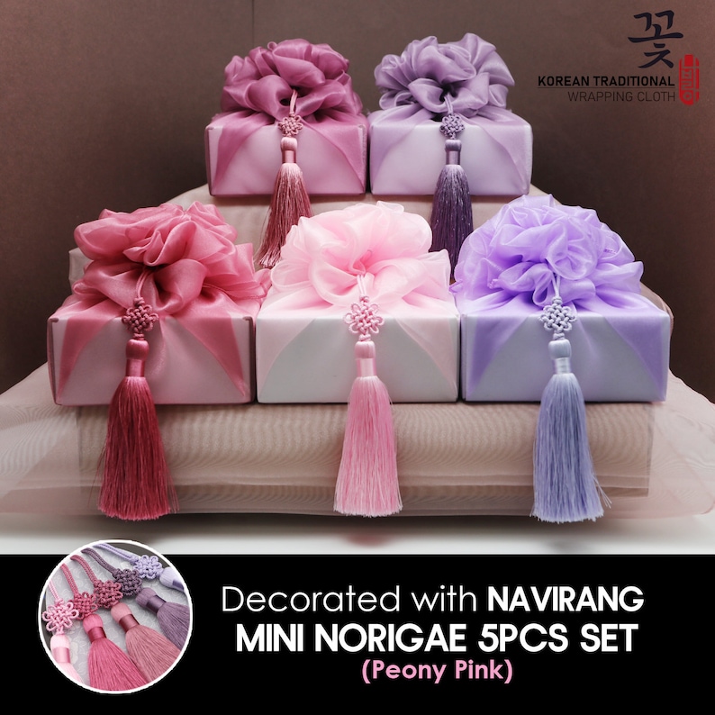 May include: Five small gift boxes wrapped in traditional Korean wrapping cloth, called "Norigae", with pink, purple, and white fabric. The boxes are decorated with a flower-shaped bow and a tassel. The text "Decorated with NAVIRANG MINI NORIGAE 5PCS SET (Peony Pink)" is at the bottom of the image.