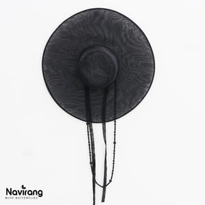 May include: A wide-brimmed black hat made of a sheer, translucent mesh fabric. The hat features a rounded crown and two long, thin black ribbons hanging down, one of which is adorned with small black beads. The text Navirang WITH BUTTERFLIES appears.