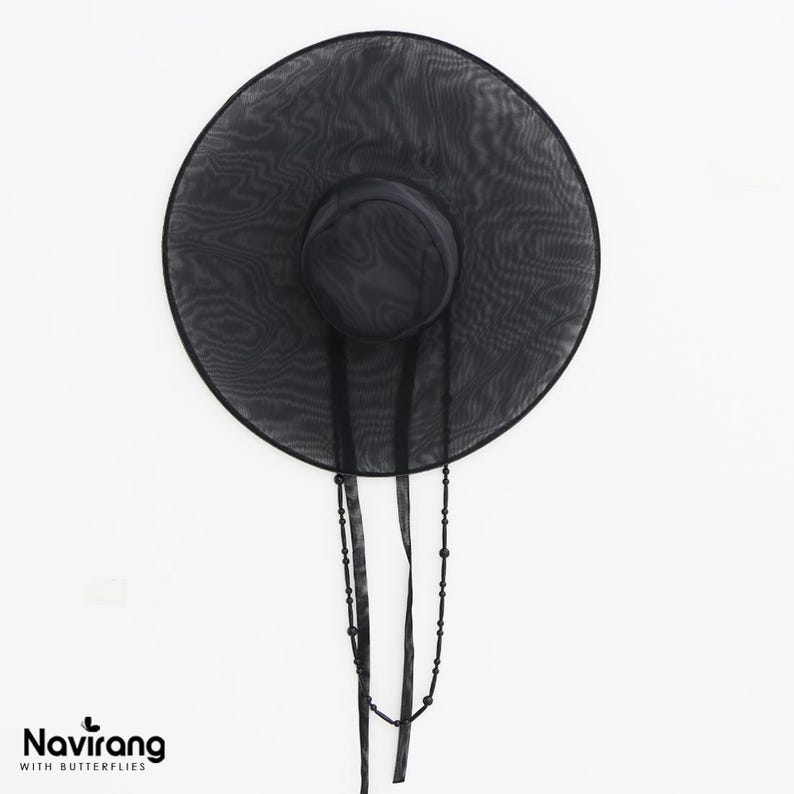May include: A wide-brimmed black hat made of a sheer, translucent mesh fabric. The hat features a rounded crown and two long, thin black ribbons hanging down, one of which is adorned with small black beads. The text Navirang WITH BUTTERFLIES appears.