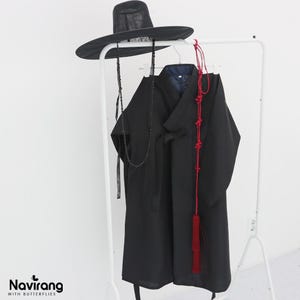 May include: A black traditional Korean-style robe with long sleeves and a wrap-front closure, paired with a wide-brimmed black hat featuring long beaded chin straps. A long red decorative cord with intricate knots and a tassel hangs over the robe.