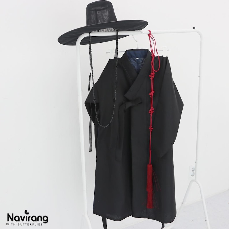 May include: A black traditional Korean-style robe with long sleeves and a wrap-front closure, paired with a wide-brimmed black hat featuring long beaded chin straps. A long red decorative cord with intricate knots and a tassel hangs over the robe.