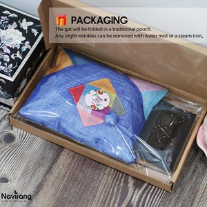 May include: A blue fabric item folded inside a clear plastic bag within a brown cardboard box. The package includes a circular sticker with floral designs and the text Thank You, alongside a black rectangular object and a small colorful patch.
