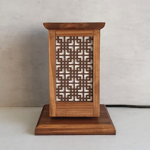 May include: A wooden table lamp with a geometric pattern on the shade. The lamp has a square base and a rectangular shade.