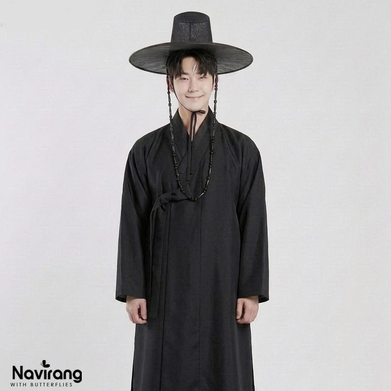 May include: A black traditional Korean hanbok robe featuring long sleeves and a wrap-around front closure with a tie. The outfit includes a wide-brimmed black hat with a beaded chin strap. The text Navirang WITH BUTTERFLIES is visible.