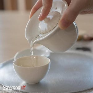 May include: A white ceramic tea set with a teapot pouring water into a small teacup. The teapot has a lid with a decorative element. The teacup is sitting on a light gray surface. The image includes the text "Navirang HANDEMADE in Korea".