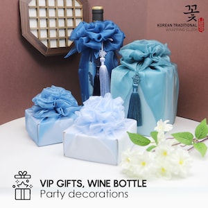 May include: Three blue and light blue gift boxes wrapped in traditional Korean wrapping cloth with a decorative knot and tassel. The boxes are arranged on a white surface with white flowers. The text "VIP GIFTS, WINE BOTTLE Party decorations" is at the bottom of the image.