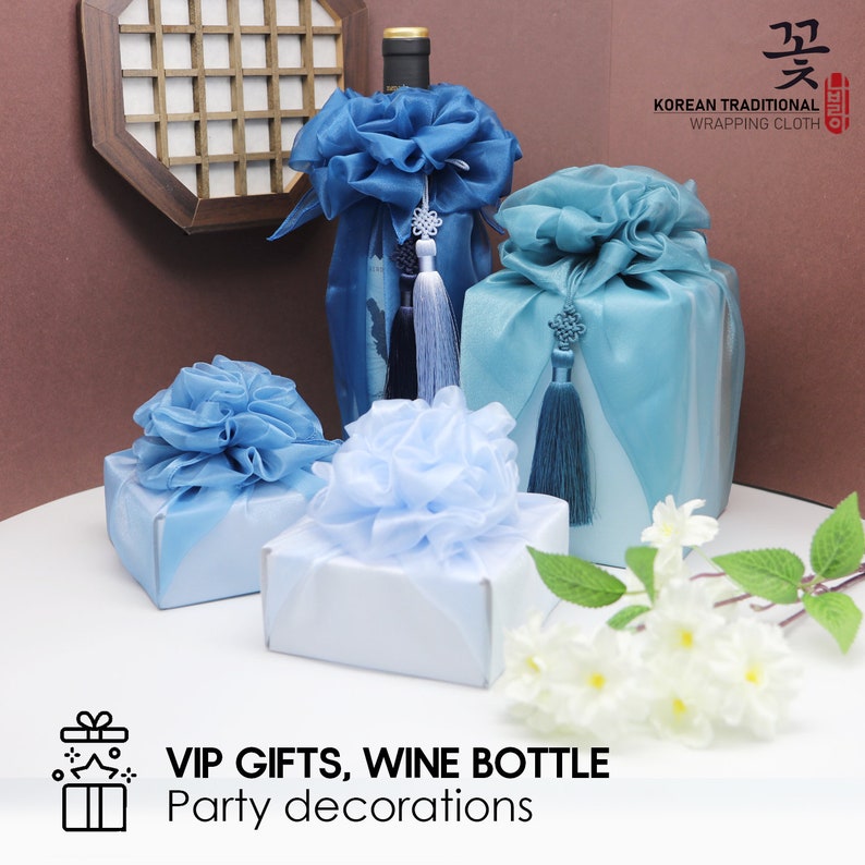 May include: Three blue and light blue gift boxes wrapped in traditional Korean wrapping cloth with a decorative knot and tassel. The boxes are arranged on a white surface with white flowers. The text "VIP GIFTS, WINE BOTTLE Party decorations" is at the bottom of the image.