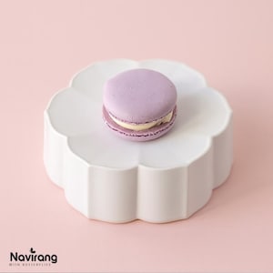 May include: A lavender macaron with a cream filling sits on a white, flower-shaped stand. The background is a pale pink. This image showcases a classic French pastry, ideal for a dessert display or afternoon tea.