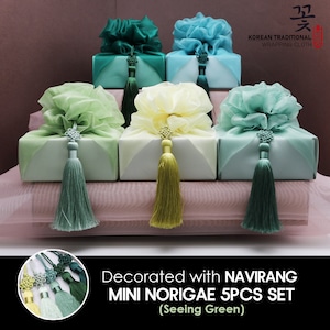 May include: Five small gift boxes wrapped in green, blue, and yellow fabric with matching tassels. The boxes are decorated with a traditional Korean knot called a "norigae". The text "Decorated with NAVIRANG MINI NORIGAE 5PCS SET (Seeing Green)" is below the boxes.