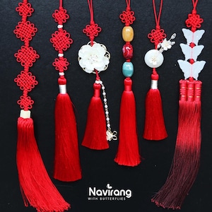 Pode incluir: Seis ornamentos decorativos suspensos com borlas de seda vermelha, cordões vermelhos com nós complexos e vários adornos, incluindo formas de flores brancas, contas de pedra coloridas e recortes de borboletas brancas. O texto Navirang WITH BUTTERFLIES é visível.