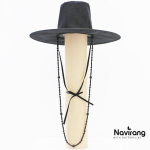 May include: A black wide-brimmed hat with a tall cylindrical crown and a long black beaded chin strap featuring small black beads and a tied ribbon detail. The text Navirang WITH BUTTERFLIES is printed in black in the bottom right corner.