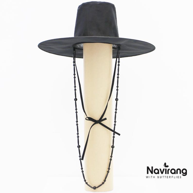 May include: A black wide-brimmed hat with a tall cylindrical crown and a long black beaded chin strap featuring small black beads and a tied ribbon detail. The text Navirang WITH BUTTERFLIES is printed in black in the bottom right corner.