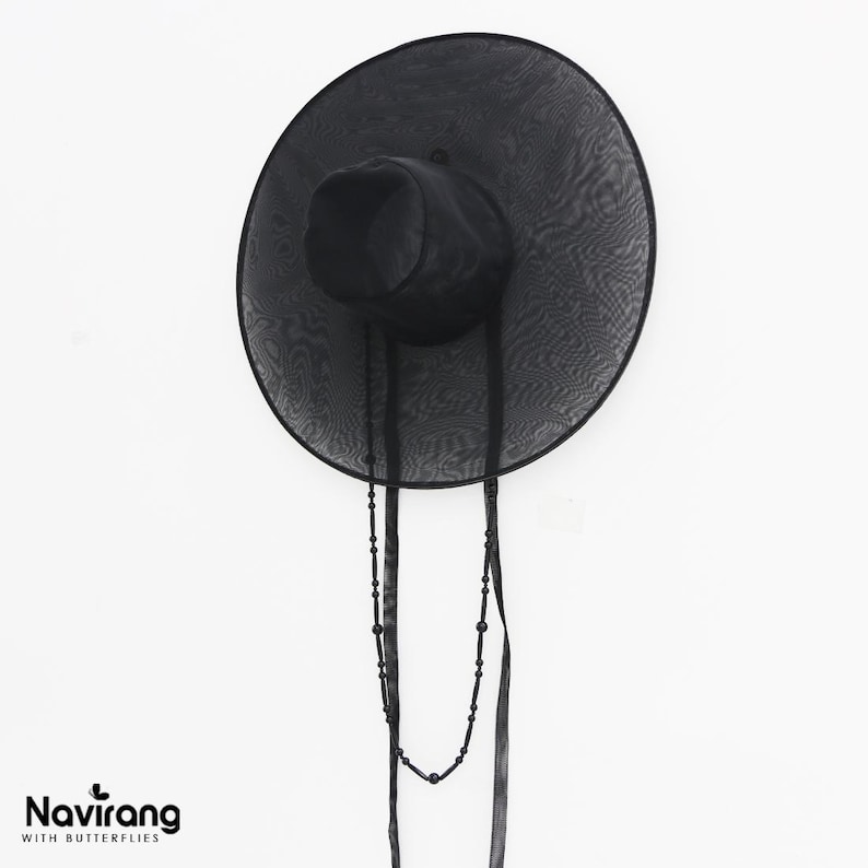 May include: A black wide-brimmed hat made of a sheer, translucent mesh material. The hat features a structured crown and a long, dangling black beaded chain accessory that hangs down from the brim. The text Navirang WITH BUTTERFLIES is visible.